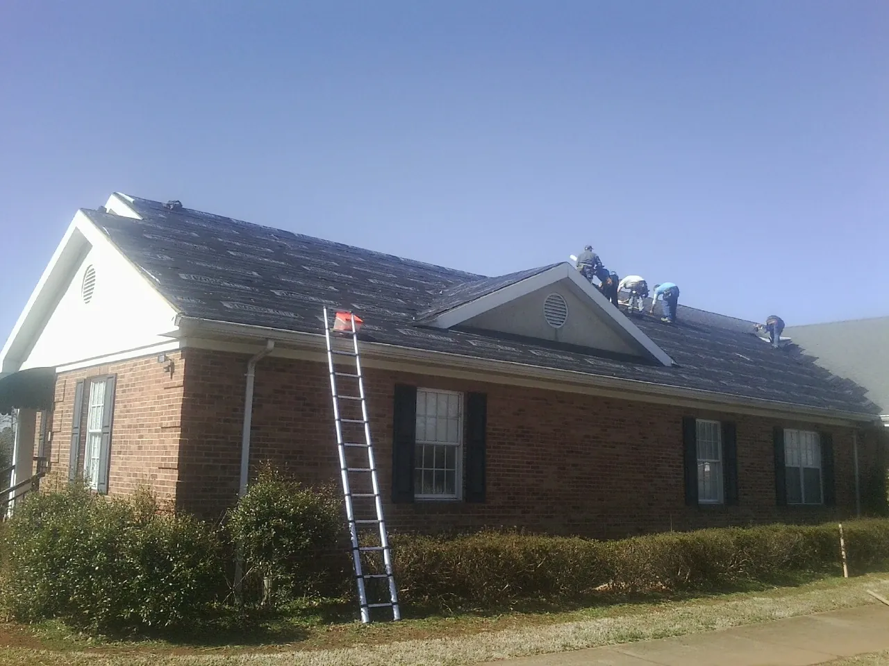 Crew tearing shingles off a brick ranch roof with a ladder set — Metal Roof Repair work by our crew in Lannon