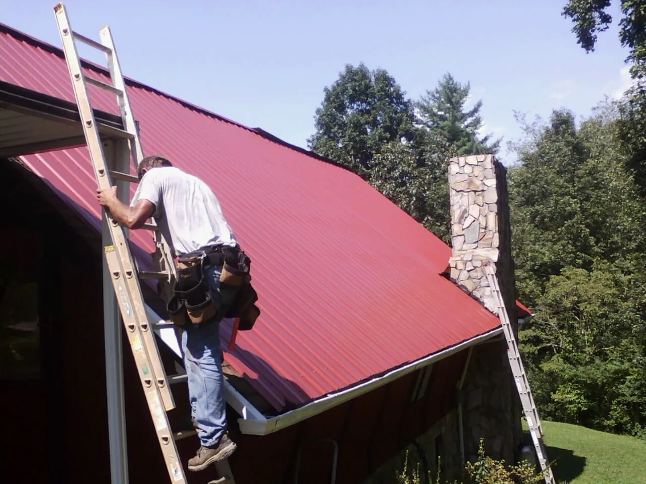 Roofer on a ladder beside a red metal roof and stone chimney — Soffit & Fascia Repair by Roofing Brothers in Lannon